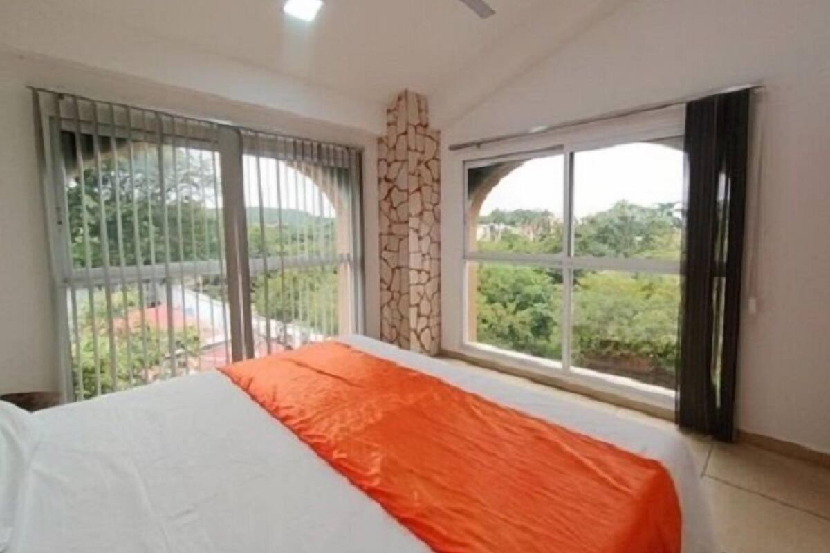 Quarto com cama, janela, ventoinha no teto e vista para o verde lá fora.