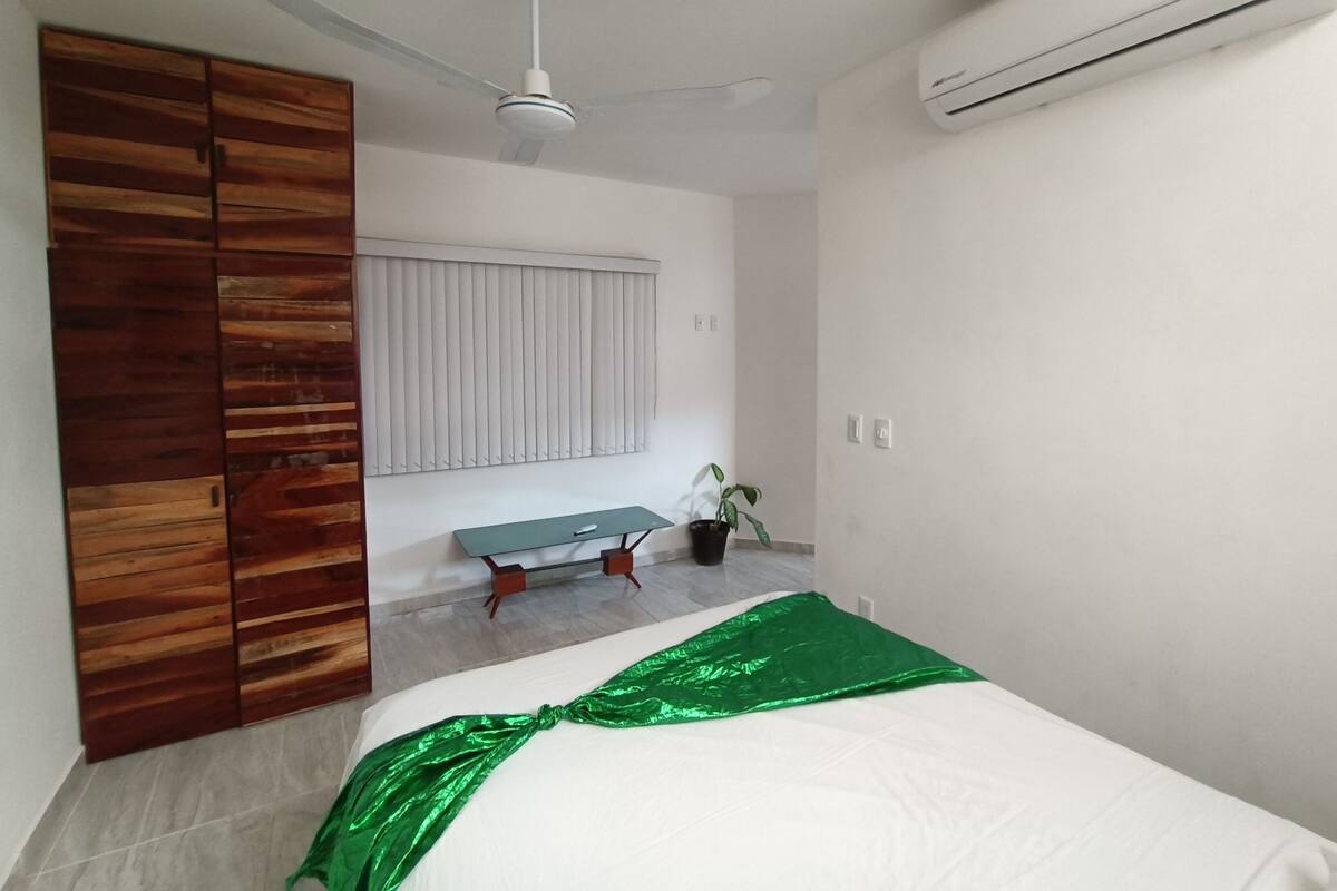 Quarto com cama, guarda-roupa de madeira, ventoinha no teto, janela com persianas, mesa pequena com planta e aparelho de ar condicionado.