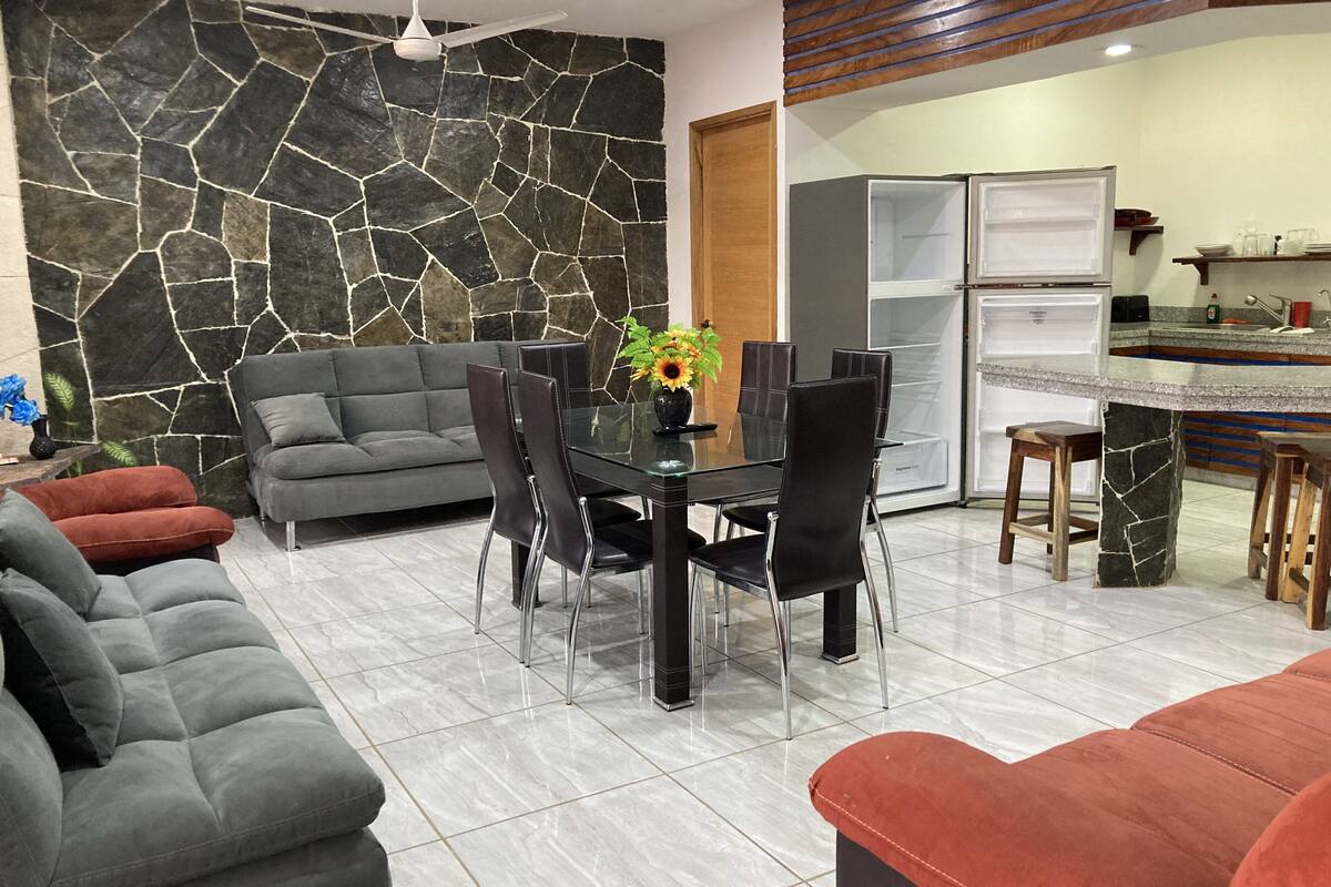 Uma sala de estar moderna com uma parede de pedra, um sofá cinzento, uma mesa de jantar com cadeiras e uma área de cozinha com frigorífico.