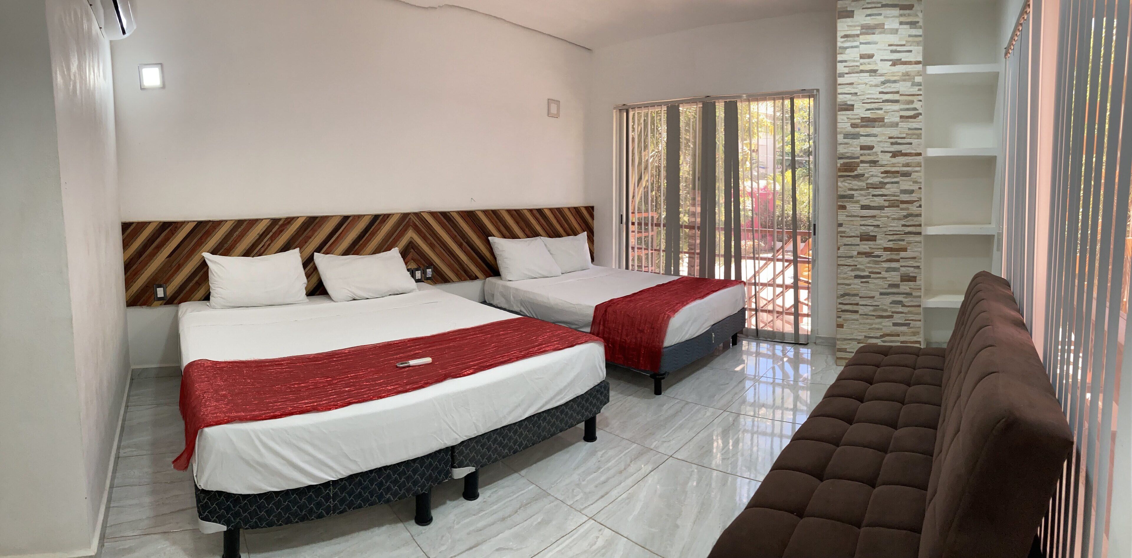 A hotel room with two beds, a brown sofa, and a view of a garden through a sliding door.