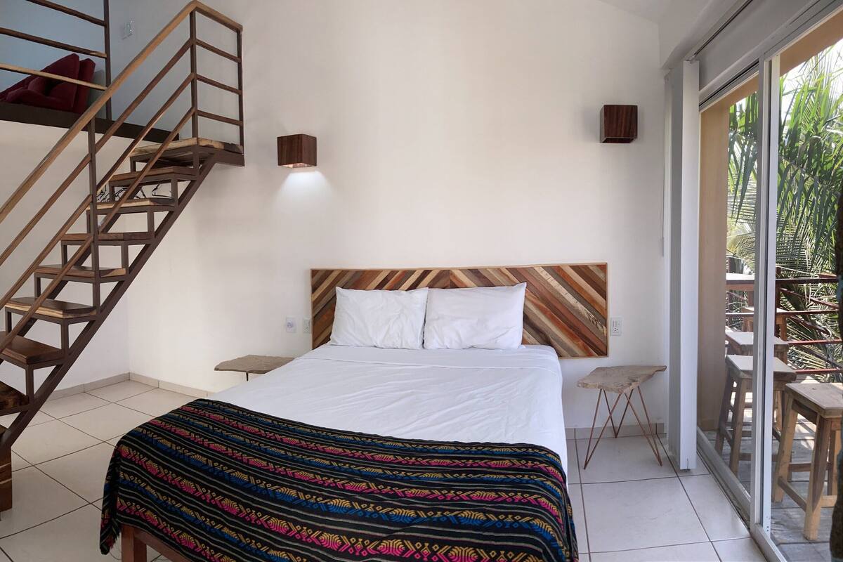 Quarto com escada de madeira, cama com roupa de cama branca, uma mesinha e vista para o exterior.