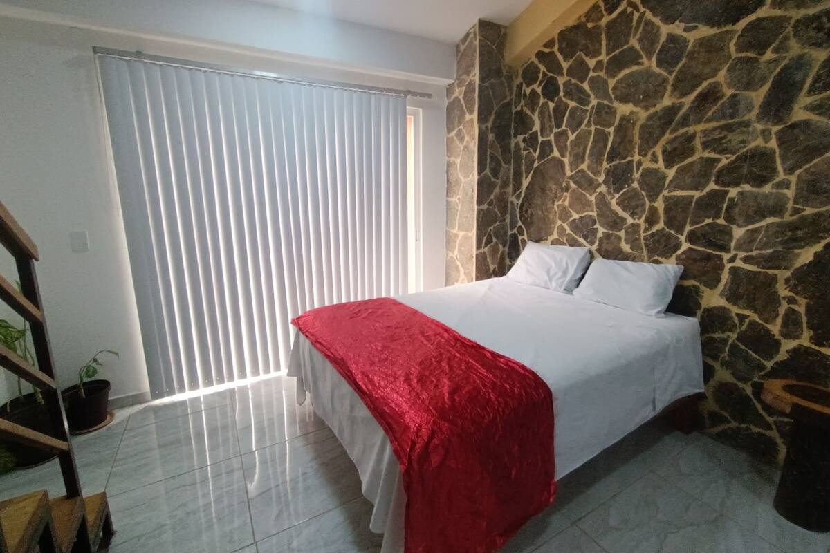 Um quarto com uma cama, parede de pedra, janela com persianas e um lance de escadas.