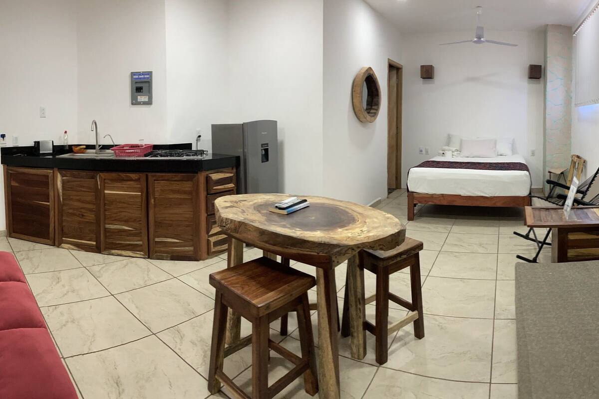 Quarto de hotel pequeno com cozinha compacta, uma cama, um sofá vermelho e uma mesa de madeira redonda.