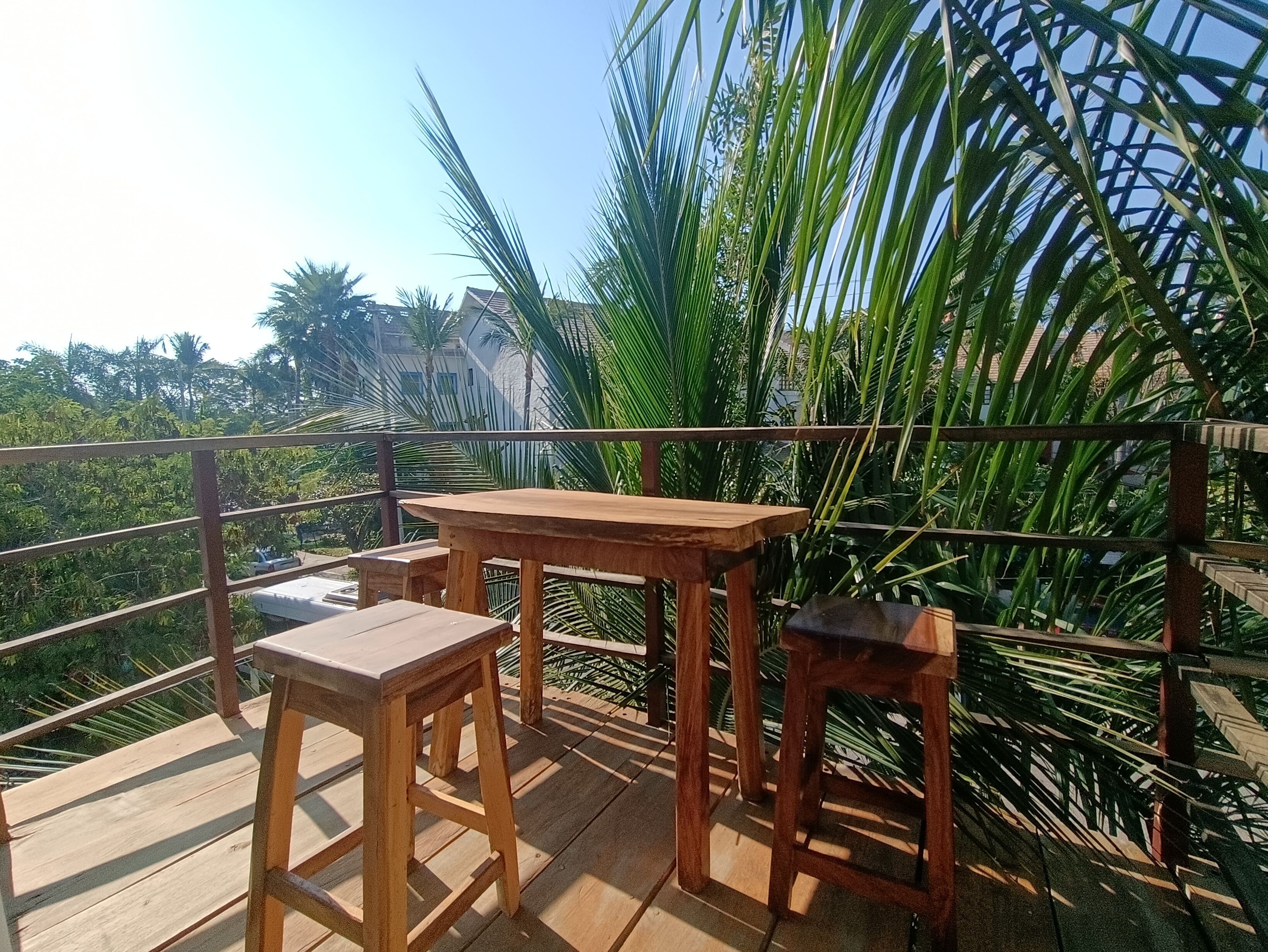 A wooden deck with a table and two stools, surrounded by lush greenery and a metal railing.