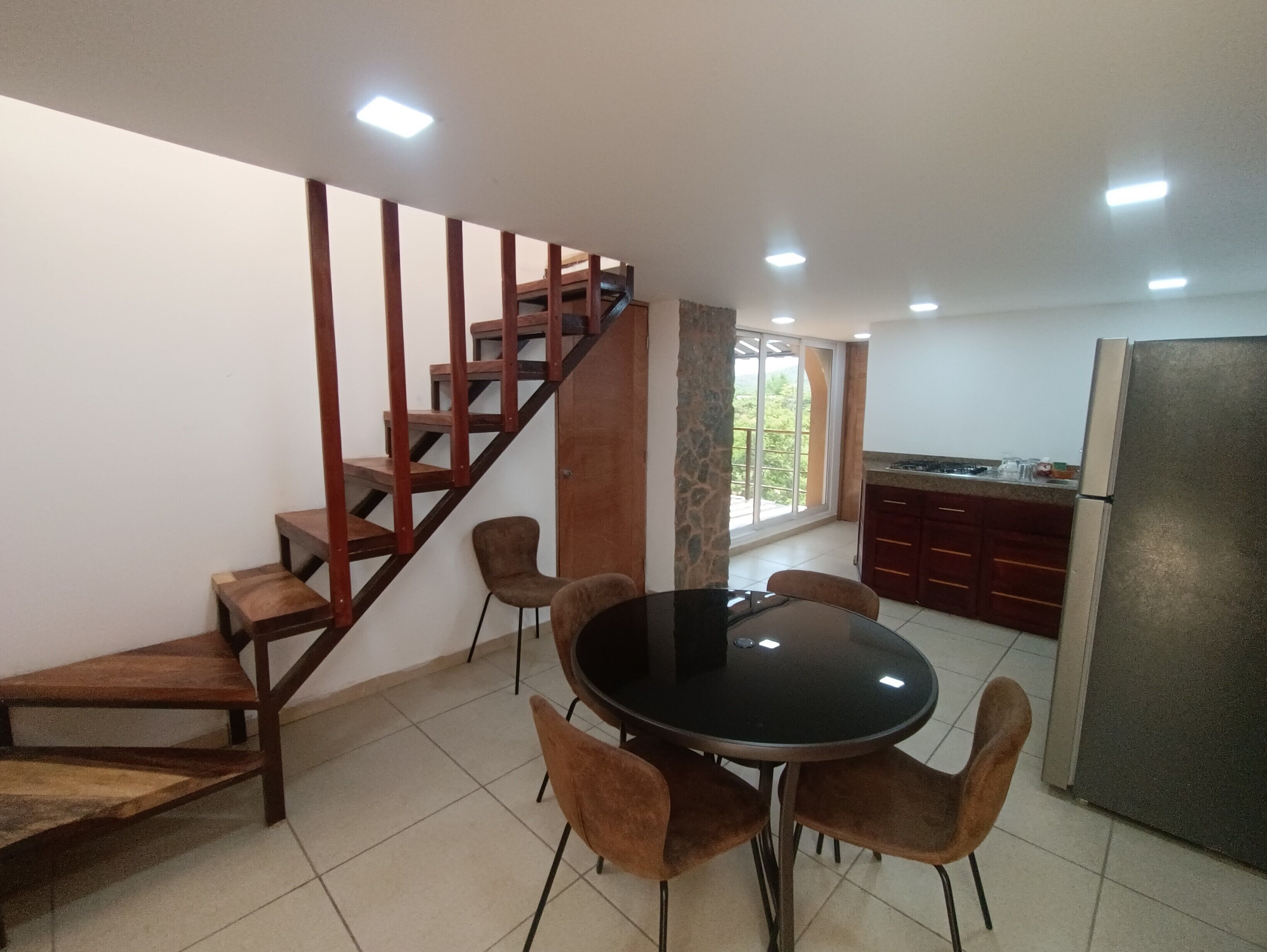 A dining area with a round table and chairs, a kitchen with a refrigerator, and a wooden staircase.