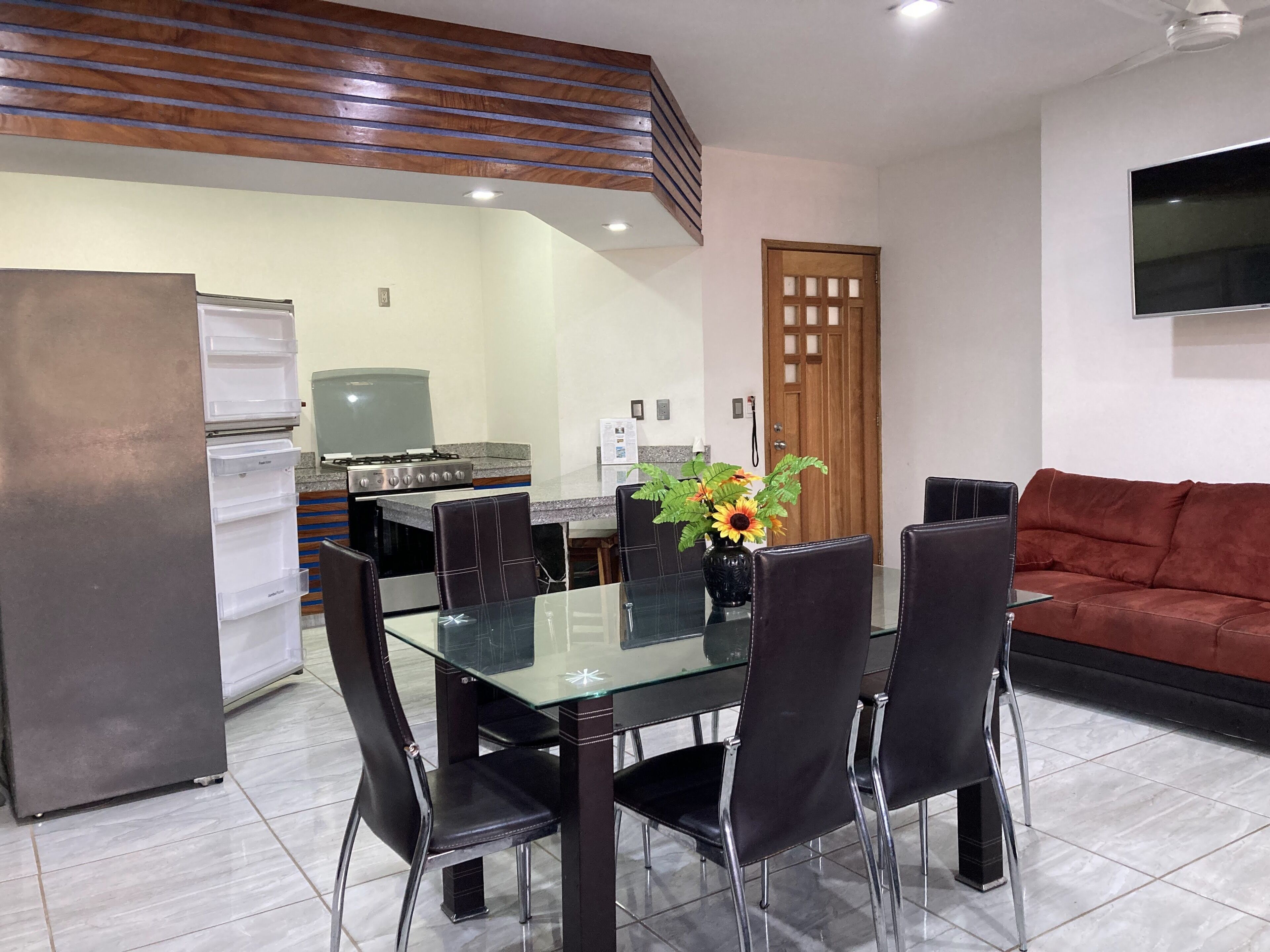A dining area with a glass table and black chairs, a kitchen with a stove and refrigerator, and a red sofa in the living area.