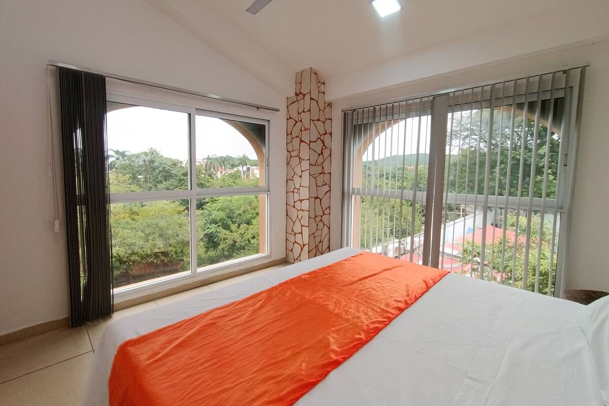 Quarto de hotel com uma cama grande, duas janelas com cortinas, um ventilador de teto e vista para o verde lá fora.
