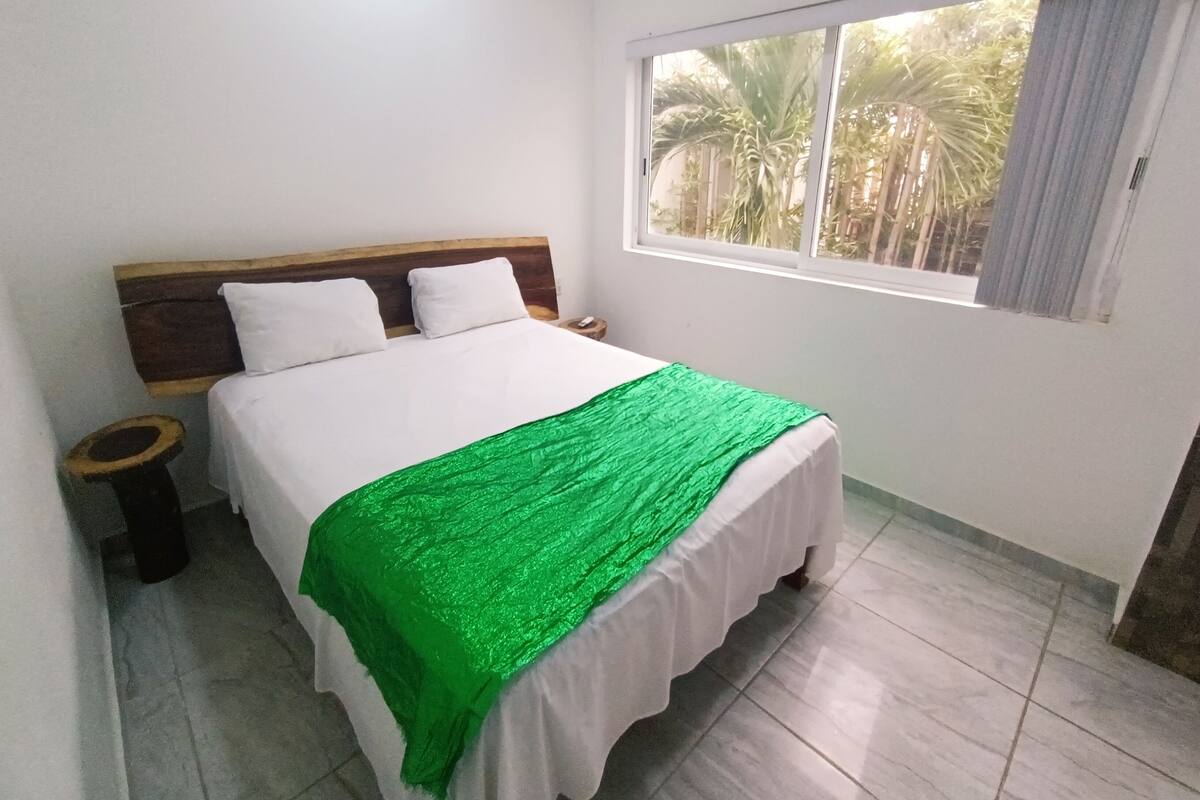 Uma cama bem arrumada com lençóis brancos e um cobertor verde, uma cabeceira de madeira, uma mesa de cabeceira com uma luminária e uma janela com vista para palmeiras.