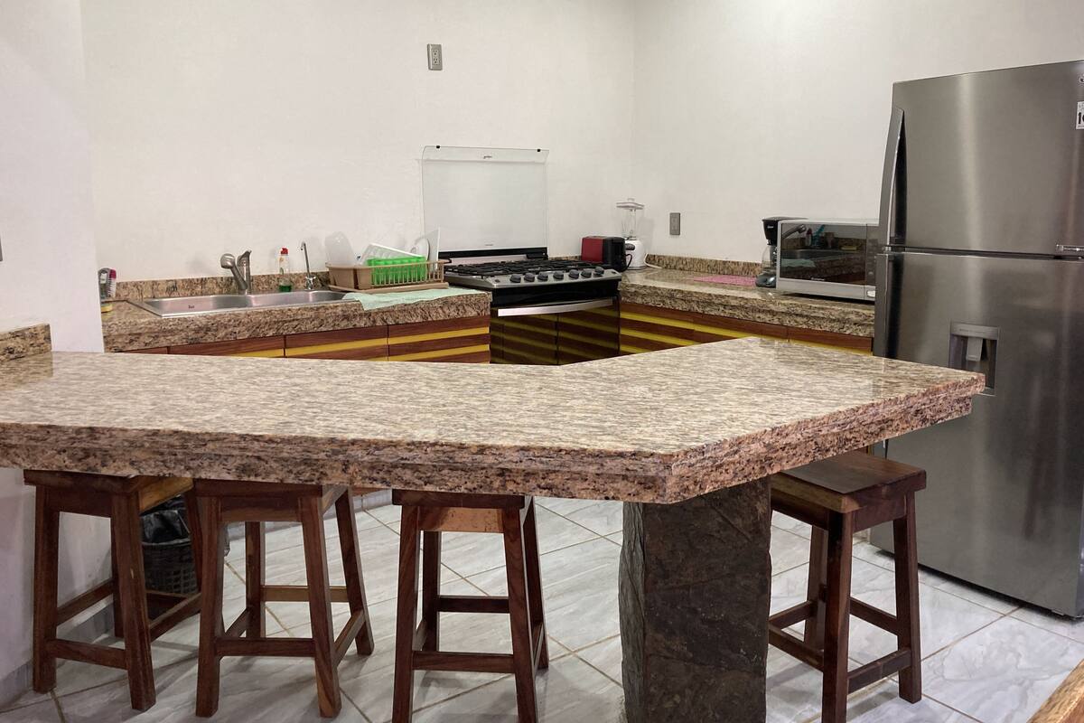 Uma cozinha com bancadas de granito, banquetas de madeira, geladeira de aço inoxidável e fogão a gás.