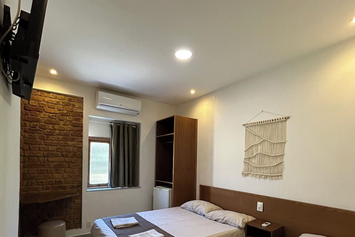 Une chambre d’hôtel avec deux lits, un mur en pierre, un climatiseur, un téléviseur fixé au mur et une décoration murale en macramé.
