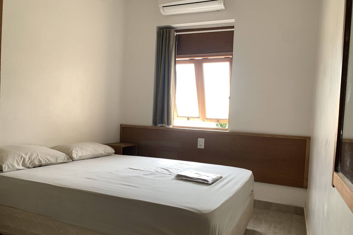 Une chambre d’hôtel comprenant un lit, une table de chevet, une fenêtre avec des rideaux et un appareil de climatisation.