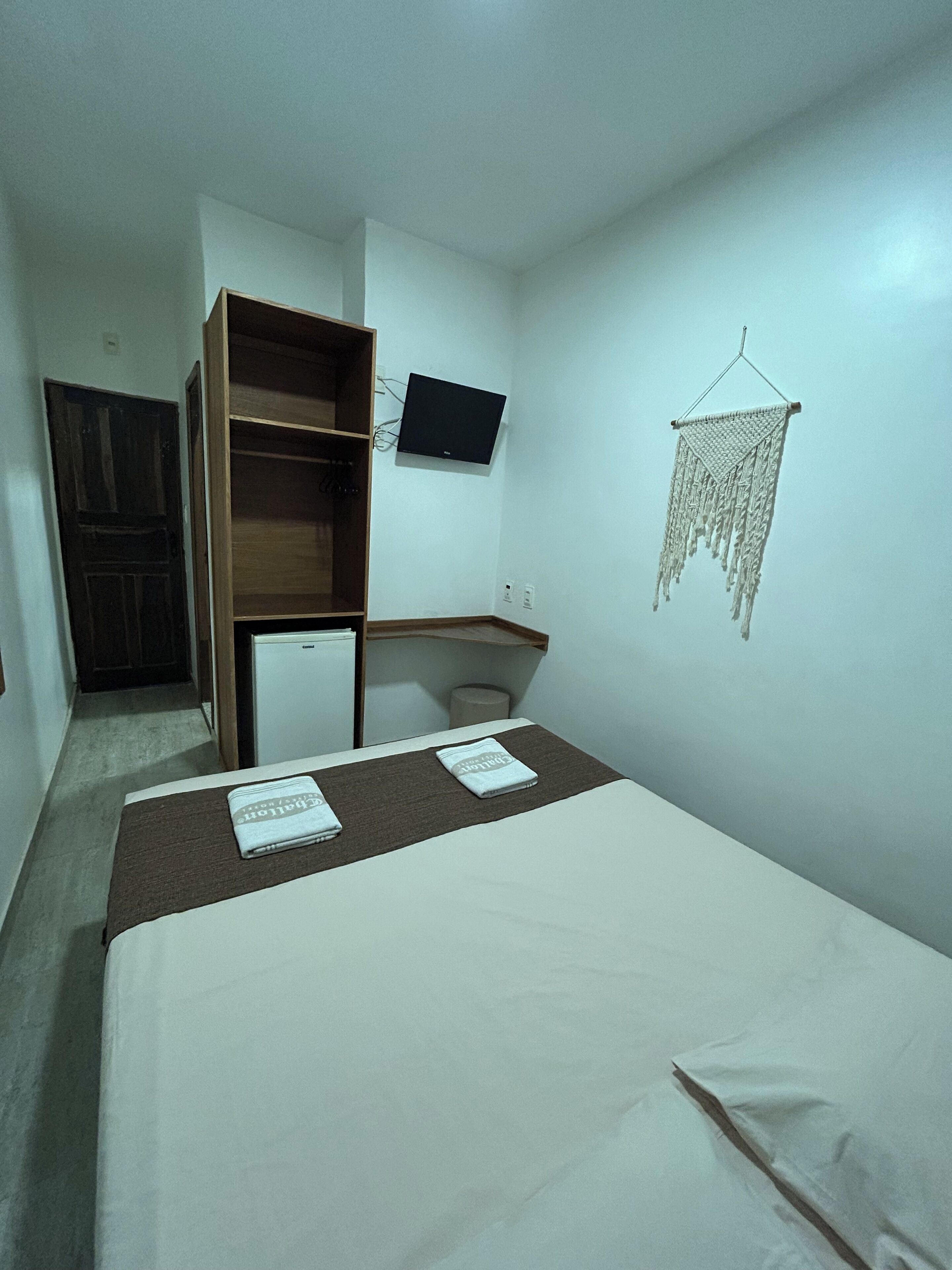 A small, sparsely furnished room with a bed, a desk, a television, and a wall hanging.