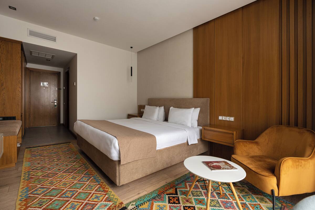 A hotel room with a bed, a small round table, a chair, and a patterned rug.
