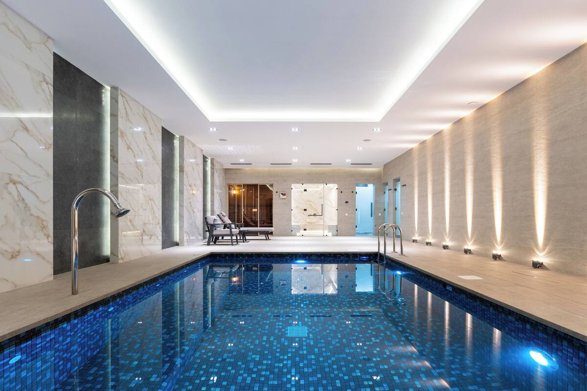 A modern indoor swimming pool with a tiled floor, a seating area with chairs, and a reception desk in the background.