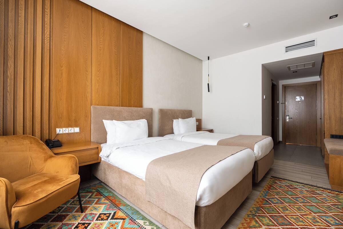 A hotel room with two beds, a wooden panel wall, a patterned rug, and a small armchair.