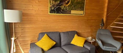 A cozy room with a grey sofa, two yellow cushions, a rocking chair, a framed picture of a bird, and a lamp.