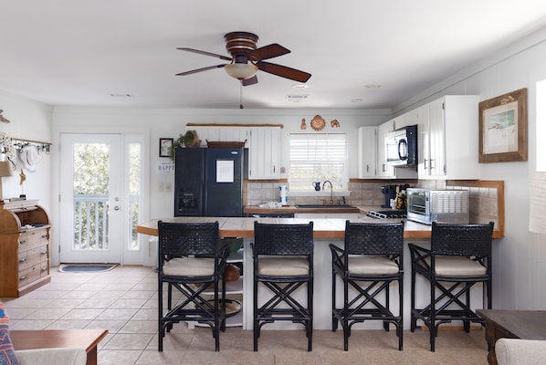 A kitchen with a central island, black wicker chairs, white cabinets, a ceiling fan, and a view of the outdoors. - Cozy, Unique - Not your typical beach rental (Gulf Shores)