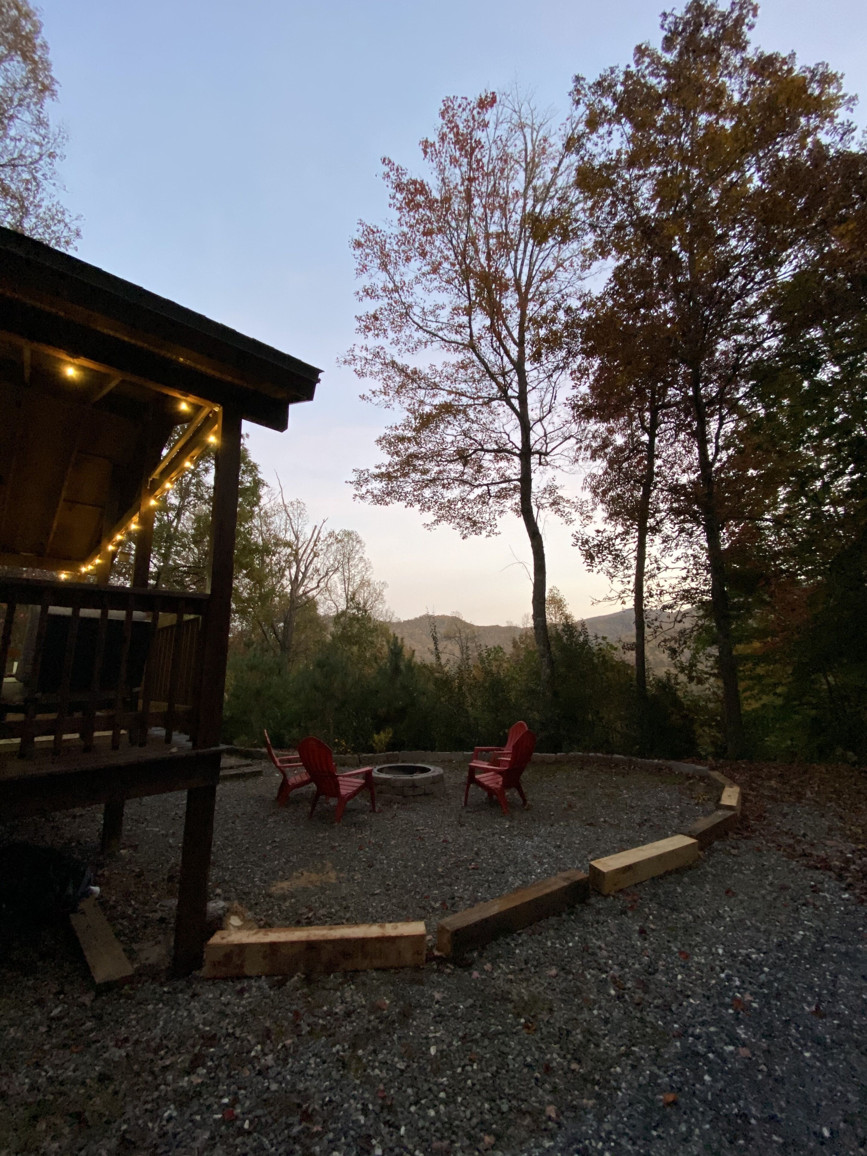 A wooden deck with string lights, two red Adirondack chairs, and a fire pit in a gravel area with trees and hills in the background.