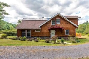 Une maison en bois à deux étages, avec un toit en pente, un porche à l’avant et une allée gravillonnée.