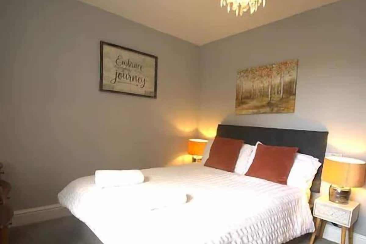 A neatly made bed with white and brown bedding, two bedside lamps, a framed inspirational quote, and a painting on the wall.