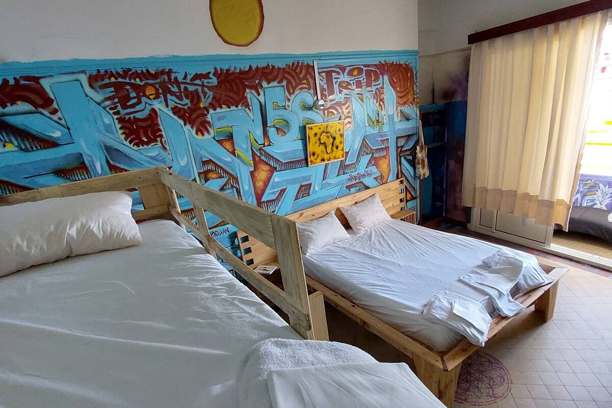 Una habitación con dos camas de madera, un mural colorido en la pared y una ventana con cortinas.