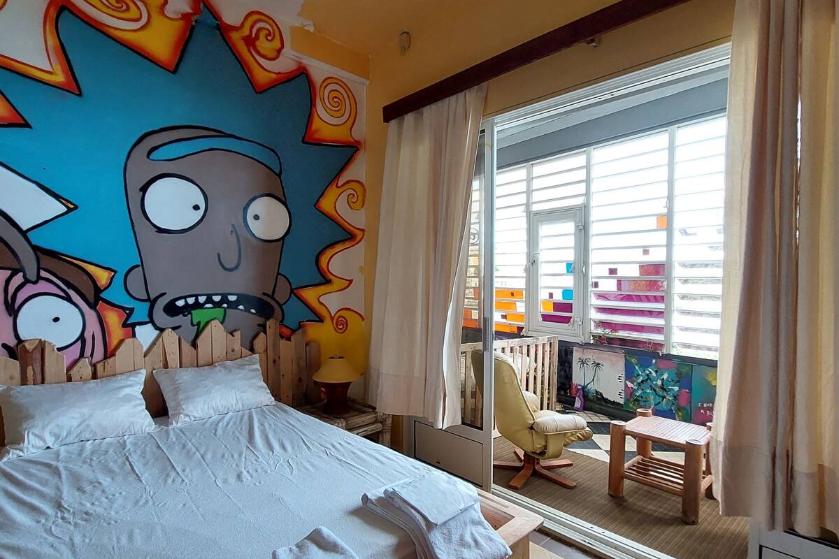 Habitación de hotel con una cama grande, un mural en la pared, una ventana con cortinas y vistas al exterior.