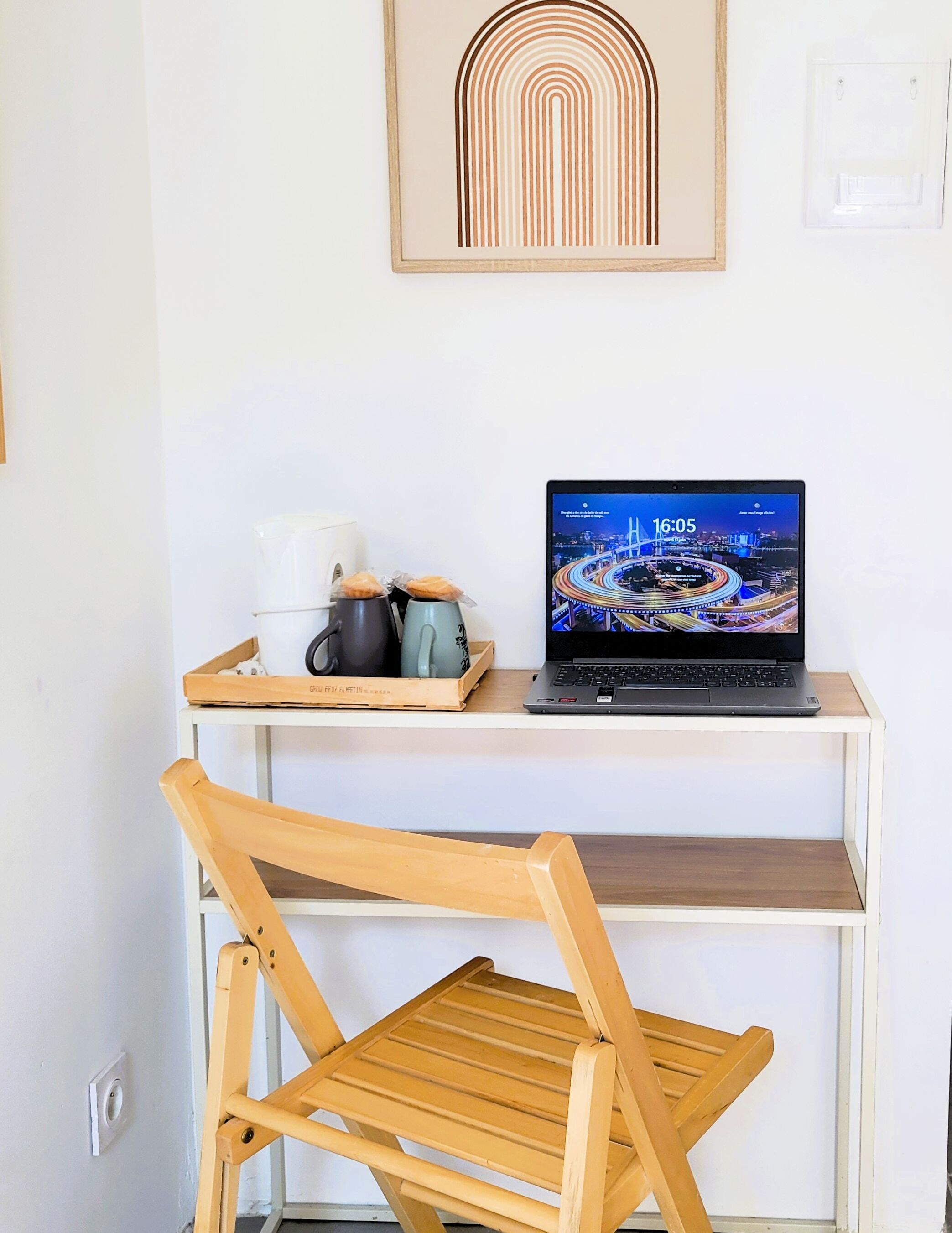 Ein Arbeitsplatz mit einem Laptop auf einem weißen Schreibtisch, einem Holzstuhl und einem gerahmten abstrakten Kunstwerk an der Wand.