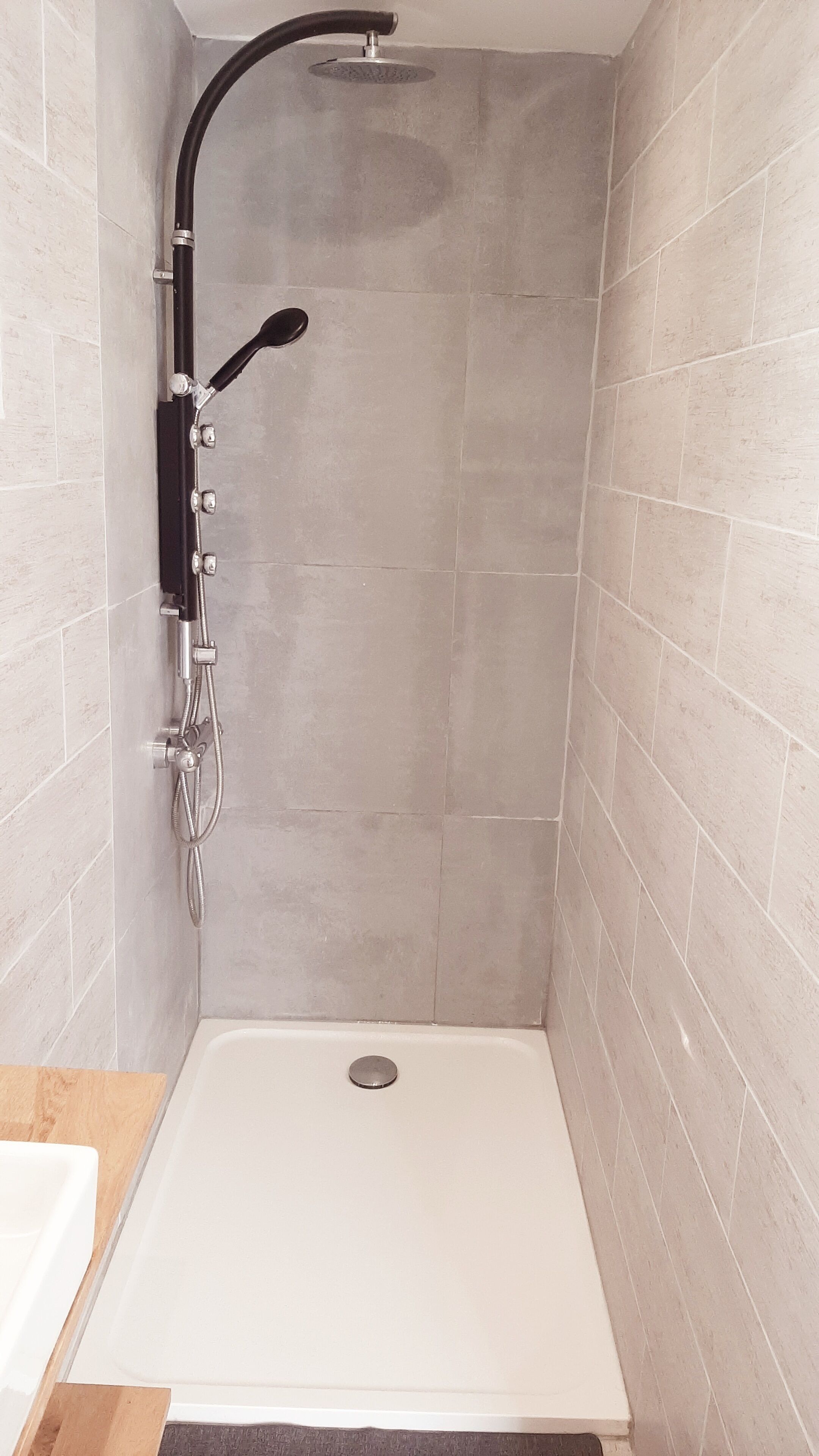 A shower with a black hose and a handheld showerhead, a white bathtub, and light grey tiled walls.