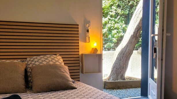 A bedroom with a bed, pillows, a bedside table with a lamp, and a view of a tree outside. - Allegria Nimes (Nîmes)