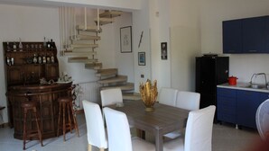A dining area with a wooden table and white chairs, a bar with stools, a staircase, and a kitchen with blue cabinets. - Portion of a villa within walking distance of Mondello. (Palermo)