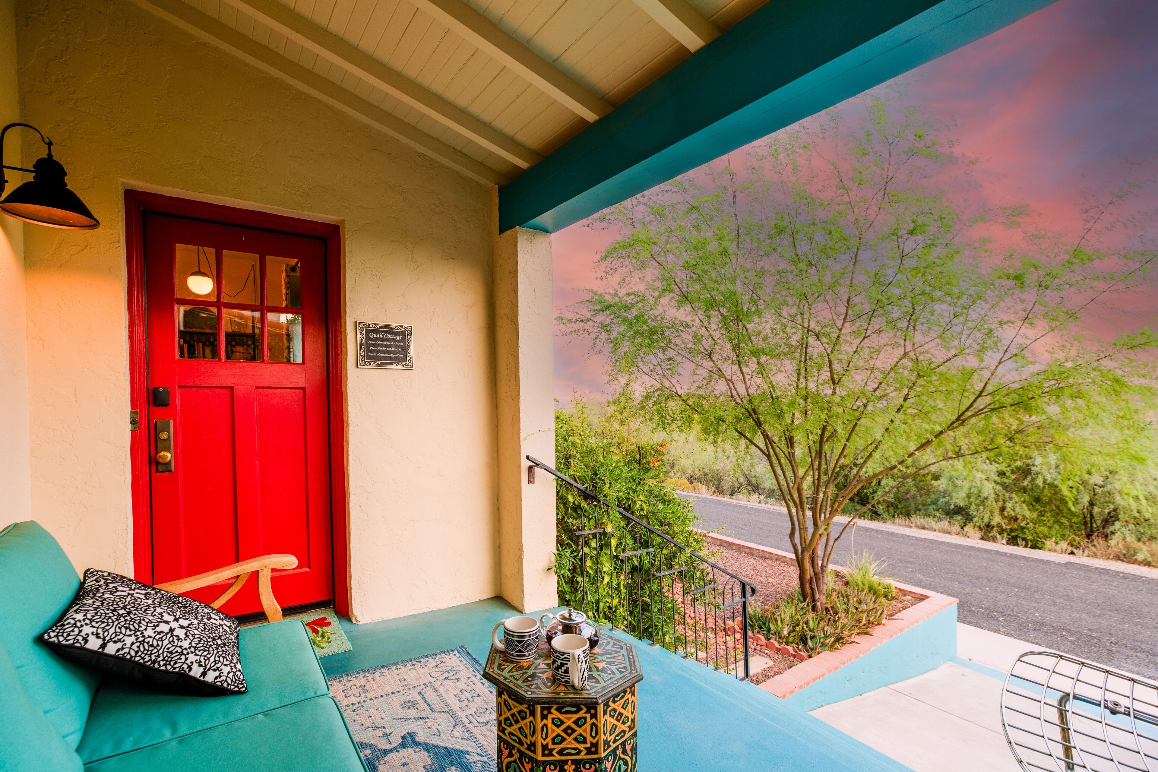 A red door, a teal sofa, and a small table with a lantern on a porch with a view of trees and a road.