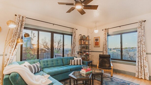 A spacious living room with a teal sofa, a black chair, and a glass coffee table. There are large windows with curtains, a bookshelf, and a ceiling fan.
