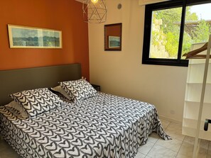 Ein Schlafzimmer mit einem Bett, zwei Kissen mit Herzmuster, einem gerahmten Bild an der Wand und einem Fenster mit Blick auf Grünflächen.