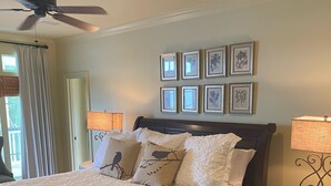 Un dormitorio con una cama grande, dos mesitas de noche, un ventilador de techo y cuadros enmarcados en la pared.