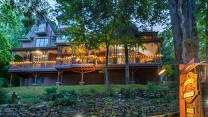 A large, two-story house with a balcony and string lights, surrounded by trees and a stone wall. - Angel At Rose Hall (Eureka Springs)