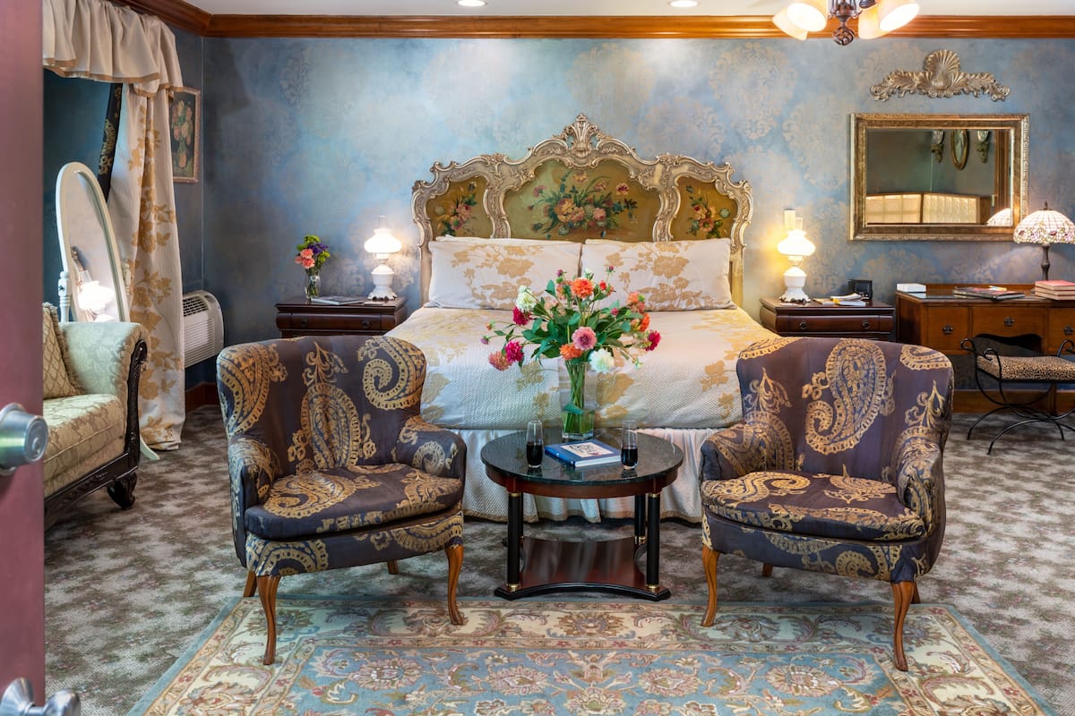 A bedroom with a large bed, two armchairs, a small table with a vase of flowers, and a nightstand with a lamp.