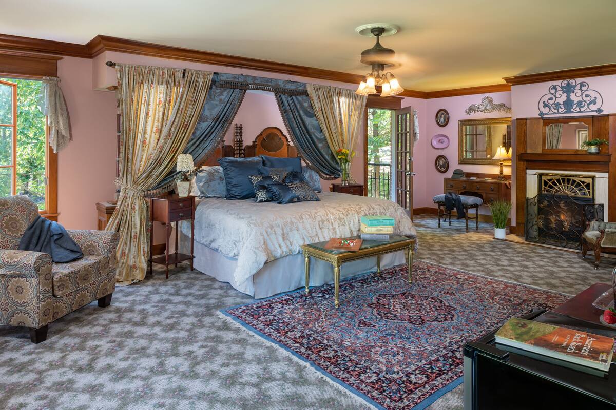 A bedroom with a large bed, a sitting area with a chair, a small table, and a fireplace.