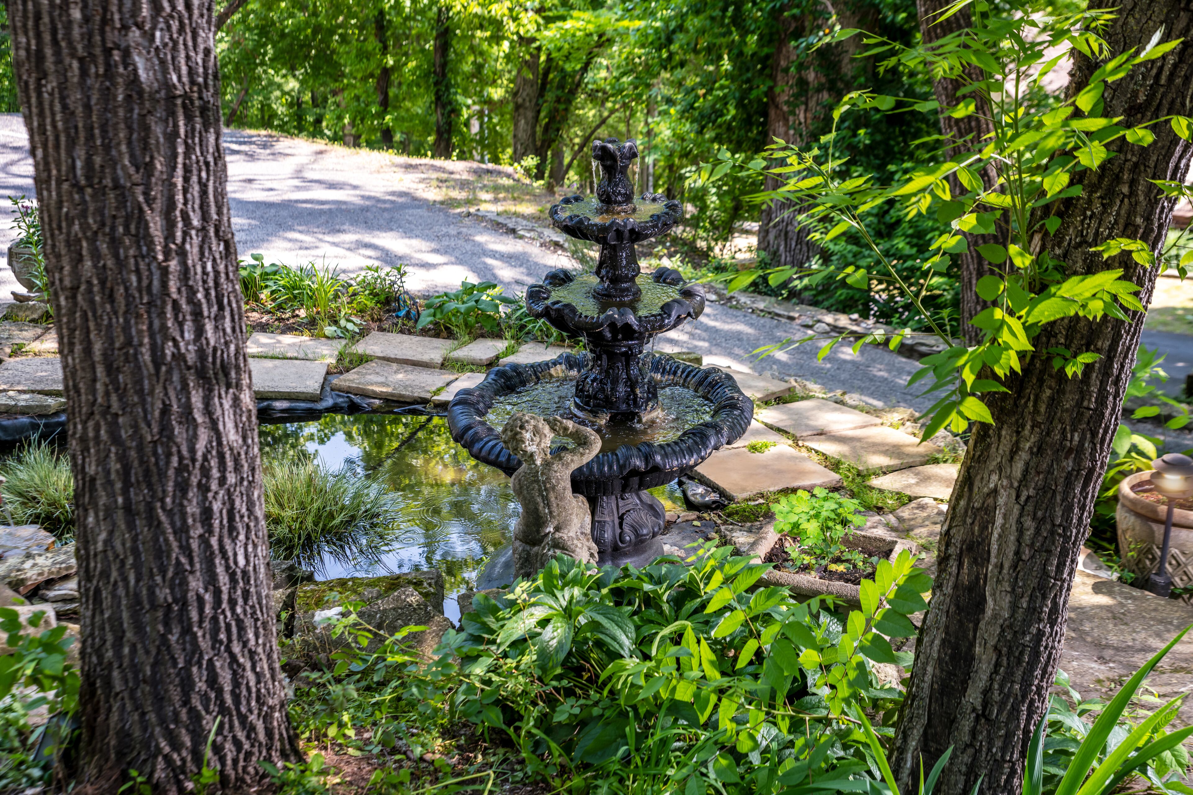 Una fuente de varios niveles en un jardín rodeado de árboles y un sendero de piedra.