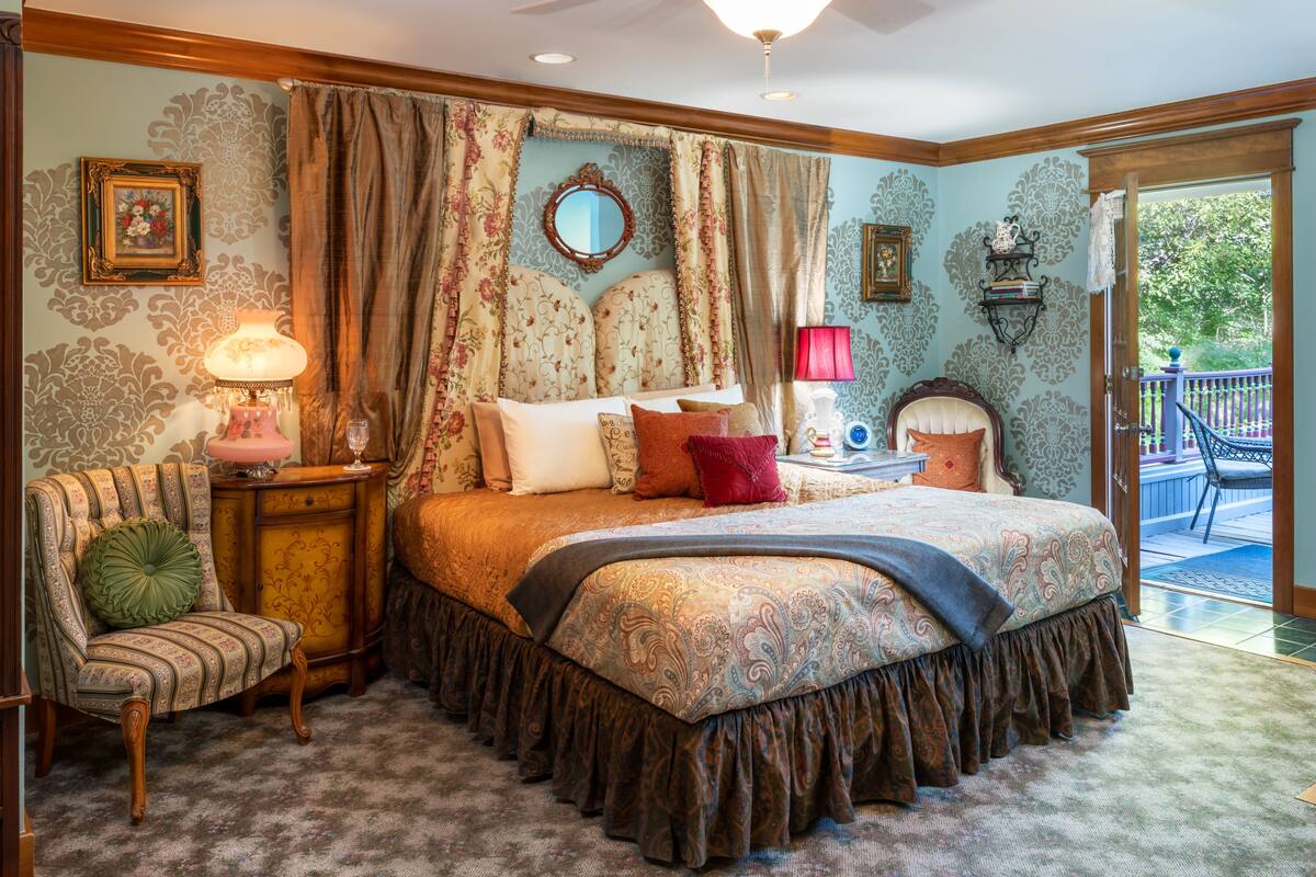 A bedroom with a large bed, a chair, a nightstand, a mirror, and a window with curtains.
