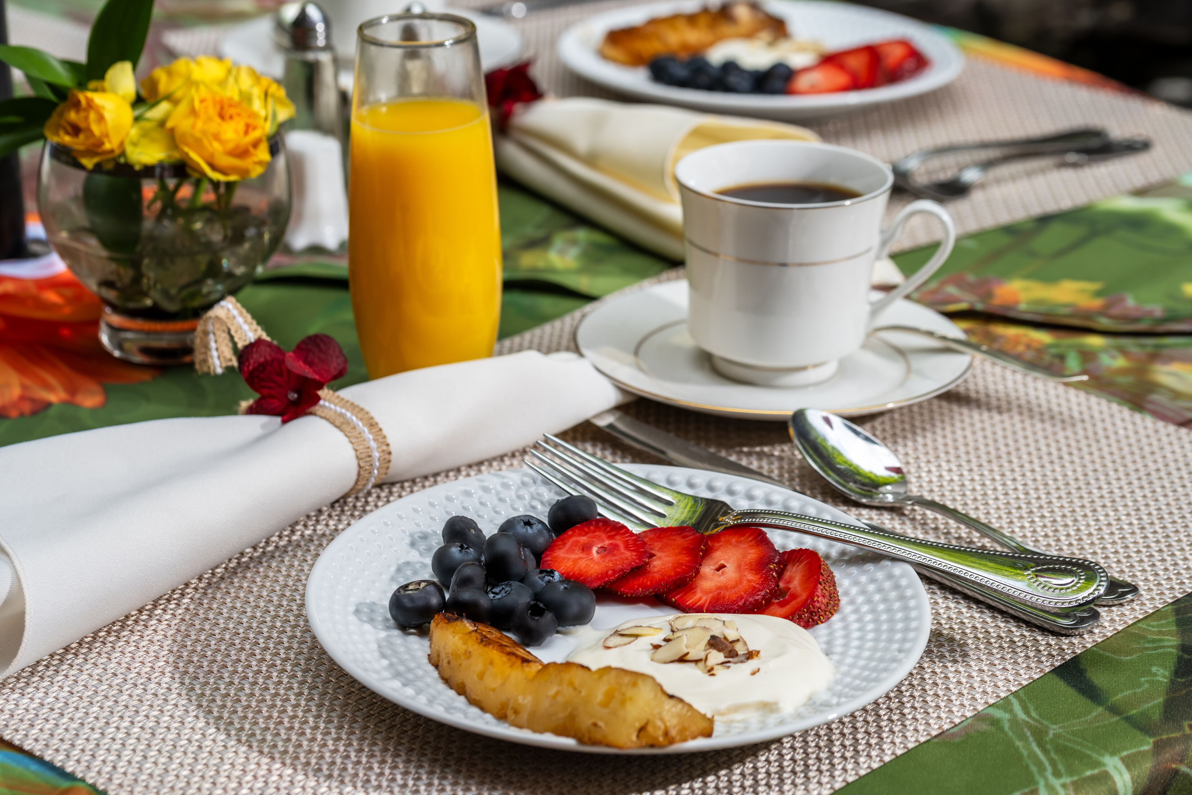 Un desayuno con un plato de yogur, arándanos, fresas y una rebanada de tostada, acompañado de un vaso de jugo de naranja y una taza de café.