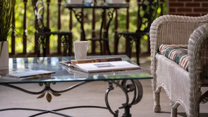 A glass-top table with a white mug, a notebook, and a pen. There is a wicker chair with a striped cushion to the right. - Angel At Rose Hall (Eureka Springs)