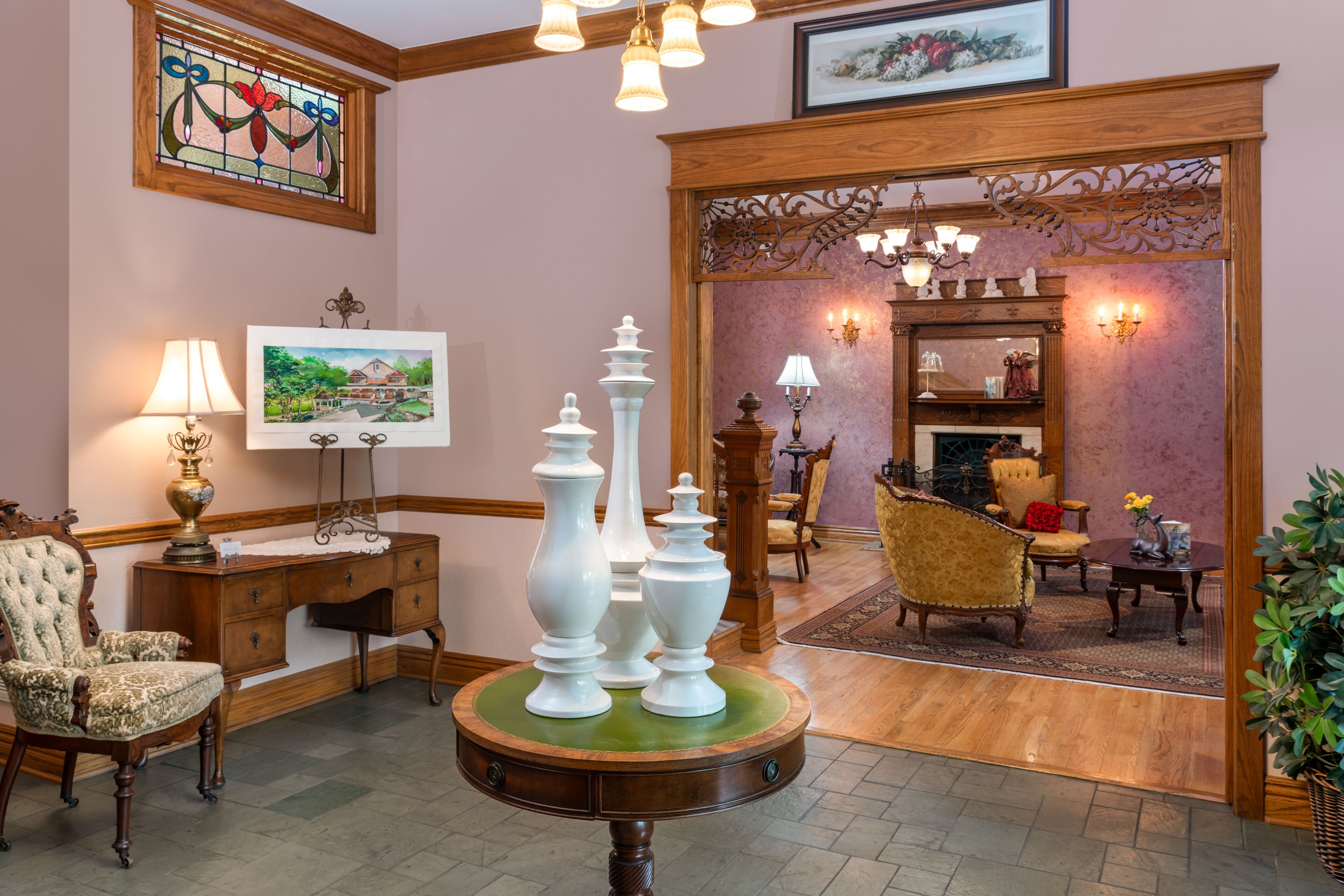 Een stijlvol ingerichte woonkamer met een houten bureau, een ronde tafel met vazen, een comfortabele fauteuil en een spiegel met wandlampen.