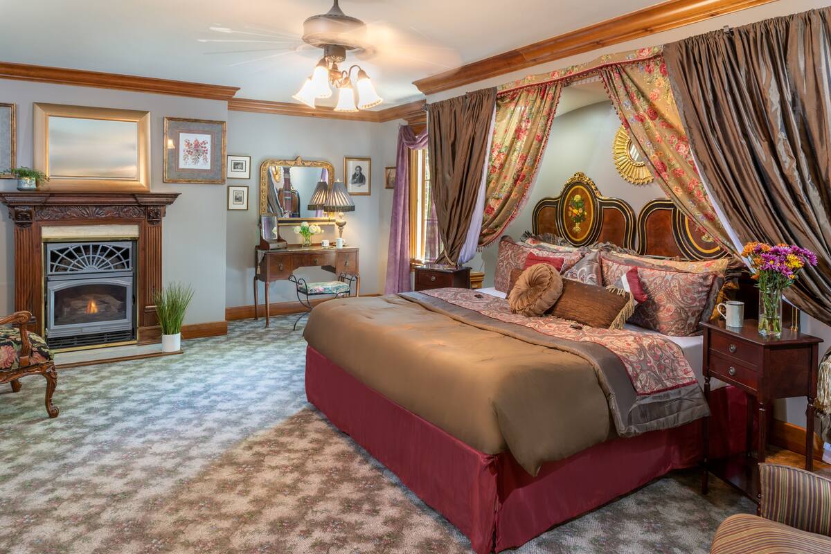 A bedroom with a large bed, a fireplace, a desk, and a chair.