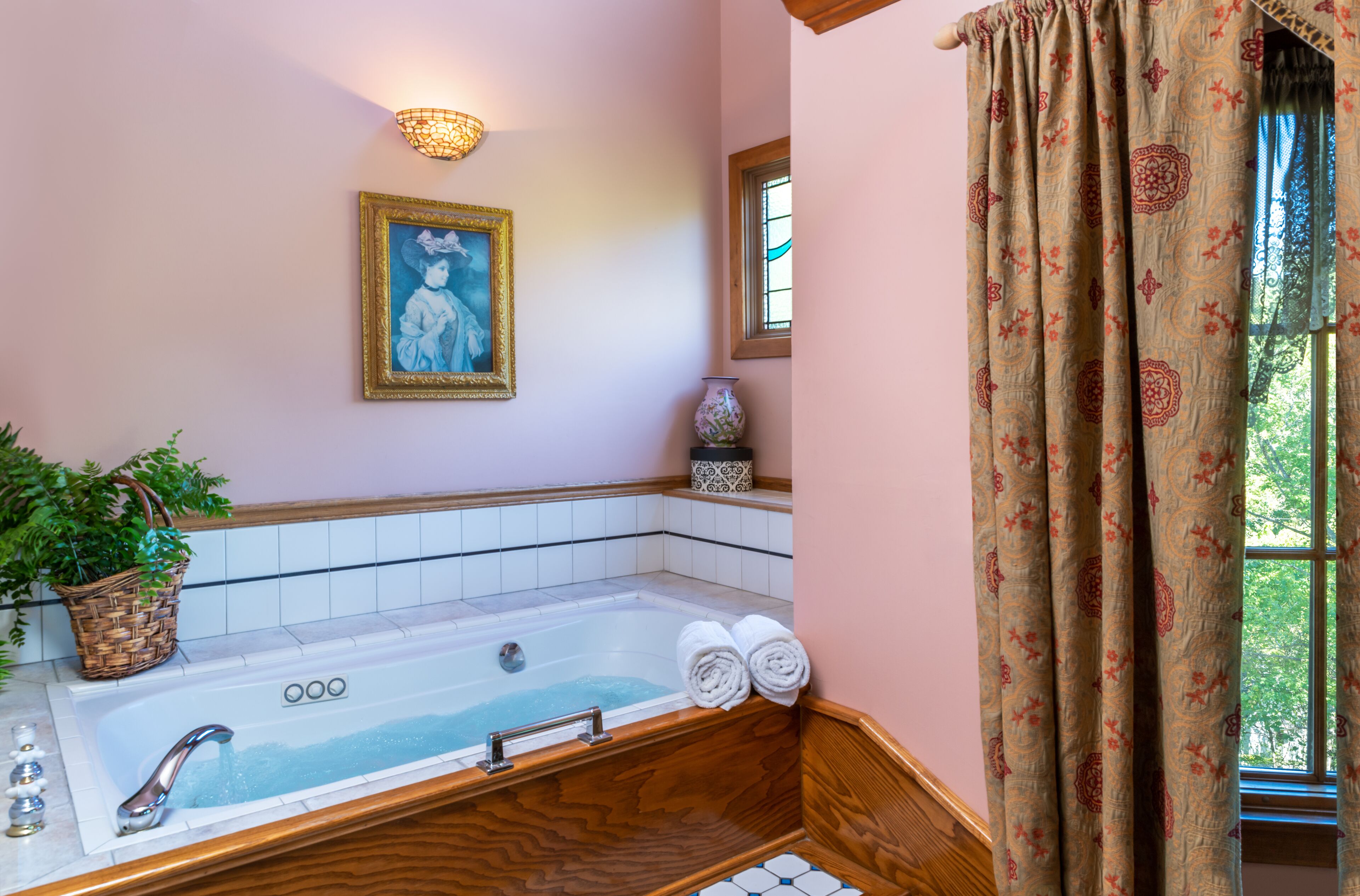 Une salle de bain avec une baignoire, un luminaire fixé au mur, un tableau encadré et une fenêtre avec des rideaux.