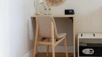 A corner room with a wooden desk, a chair, a small shelf with a radio, a decorative mirror, and a wall-mounted rack.