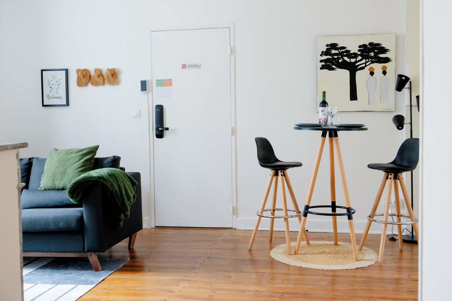 Un salon moderne avec un canapé bleu foncé, un plaid vert et un plancher en bois. On y trouve une porte blanche avec une serrure et un petit tableau encadré accroché au mur. Une table ronde, accompagnée de deux chaises noires, est placée près de la porte.
