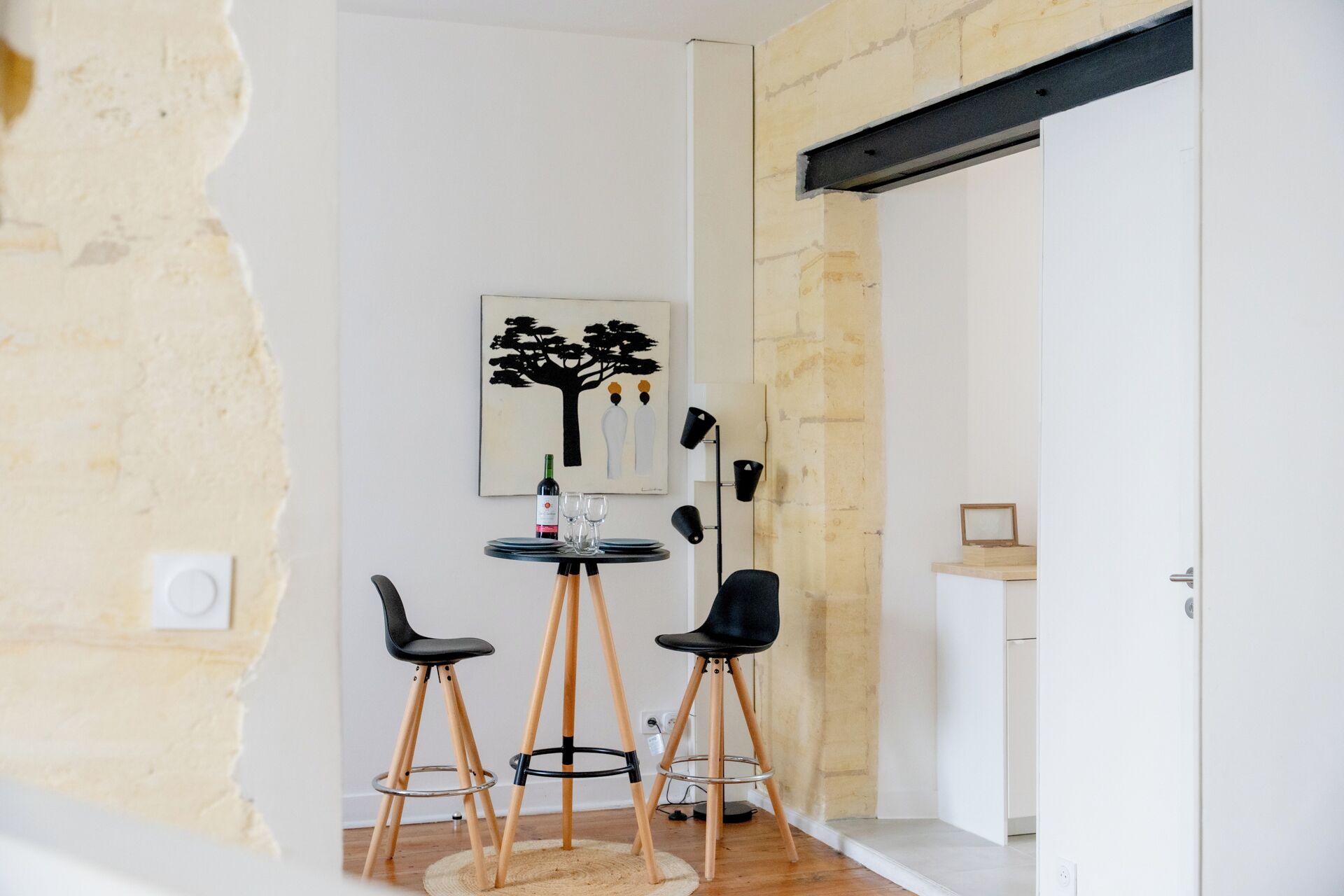 Uma área de refeições com uma mesa posta para dois, duas cadeiras pretas, um quadro de uma árvore e uma garrafa de vinho.