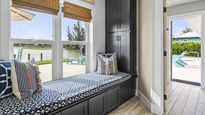 Una habitación iluminada por el sol, con un banco empotrado, ventanas amplias con persianas y vistas a una piscina y vegetación.