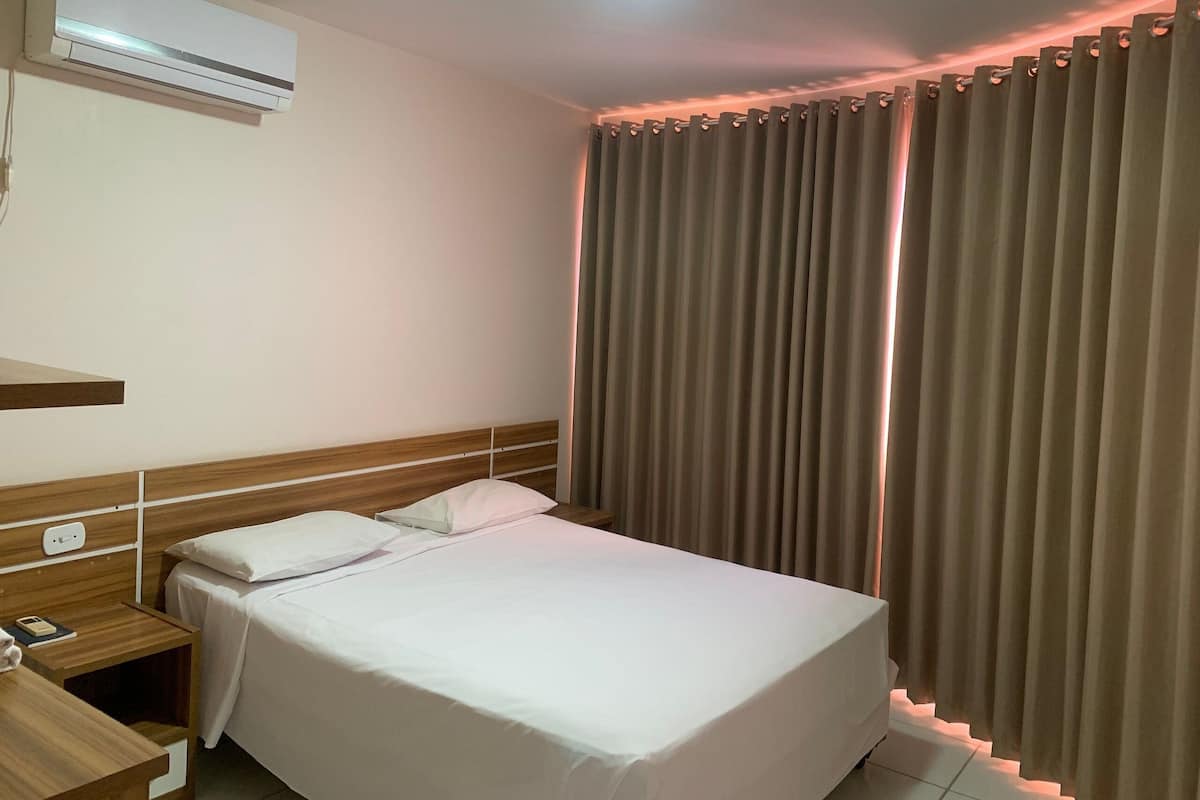 A hotel room with a bed, a bedside table, an air conditioning unit, and a window with curtains.