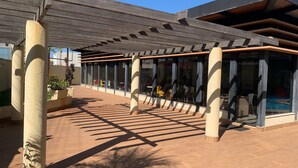 Un patio exterior con zona de estar cubierta, ventanales grandes y una piscina visible a través del vidrio.